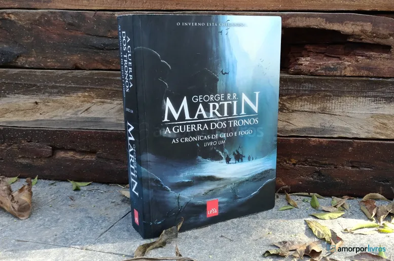 Capa do livro 'A Guerra dos Tronos' de George R. R. Martin, exibido ao ar livre com um fundo de madeira e folhas caídas